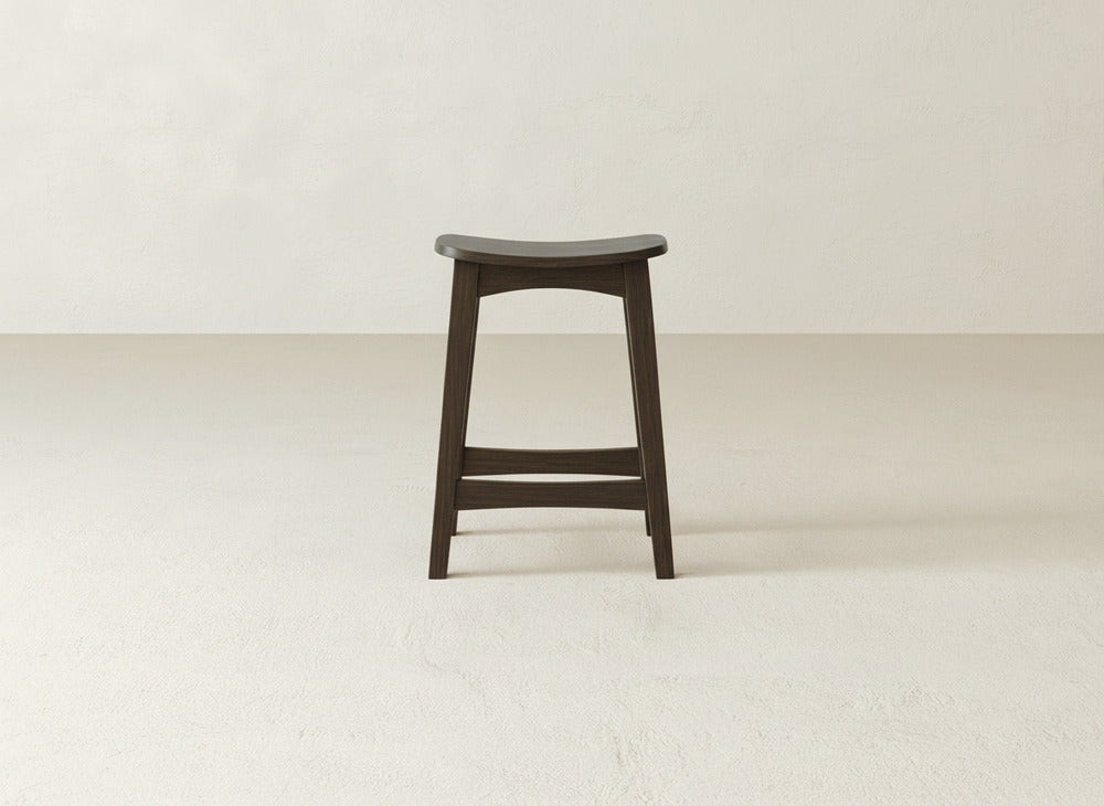The Weston Stool by James & James, featuring dark maple hardwood, a contoured seat, and natural wood grain, is centered in a minimalist room with light walls and floors.