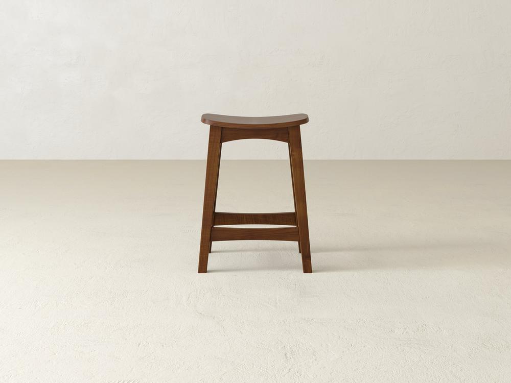 A Weston Stool by James & James, crafted from durable maple hardwood, sits with its contoured seat in a minimalist room. The light floor and plain, textured walls highlight its simple design as it stands confidently facing forward.