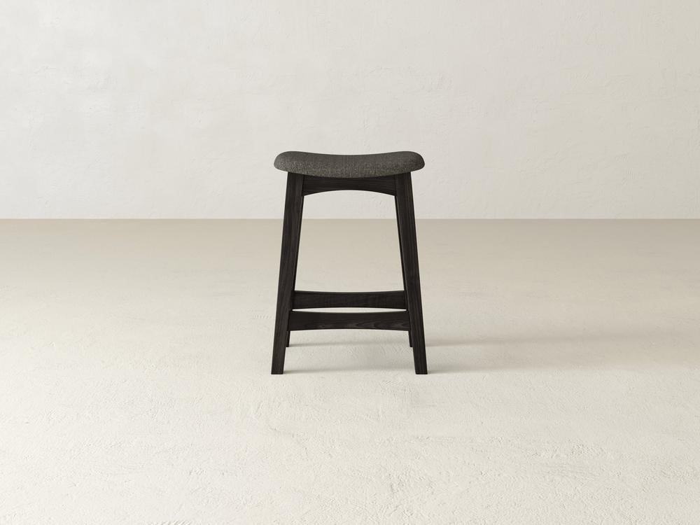 The Weston Upholstered Stool by James & James is a tall, minimalist design with a dark wooden seat made from maple hardwood. It features four legs and no backrest, standing on a beige floor against a light gray wall.
