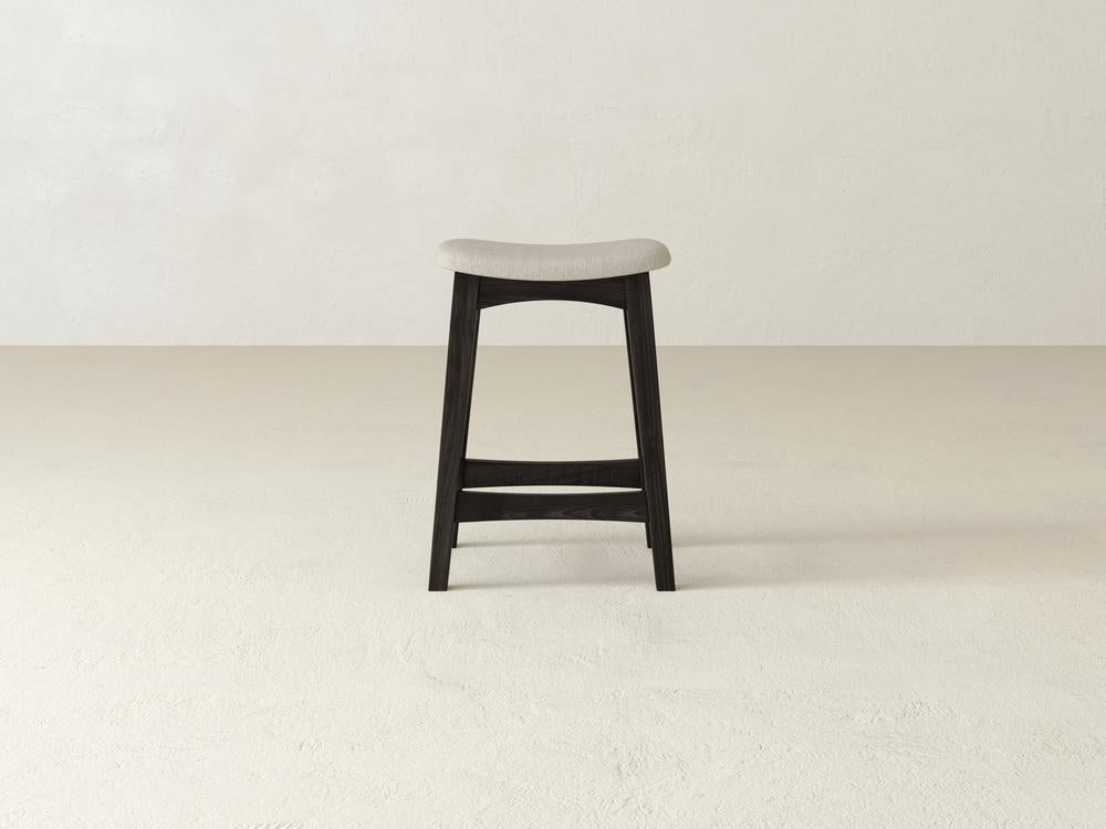 The Weston Upholstered Stool by James & James features a light grey contoured seat, crafted from durable maple hardwood. It stands on a plain, light-colored floor against an off-white textured wall and boasts a simple, modern design with a dark wooden frame.