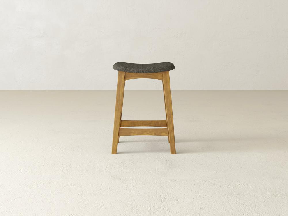 The Weston Upholstered Stool by James & James, made of maple hardwood with a dark cushion, is centered in a plain, light-colored room. This streamlined stool includes a footrest on its lower legs and an elegantly contoured seat for added comfort.