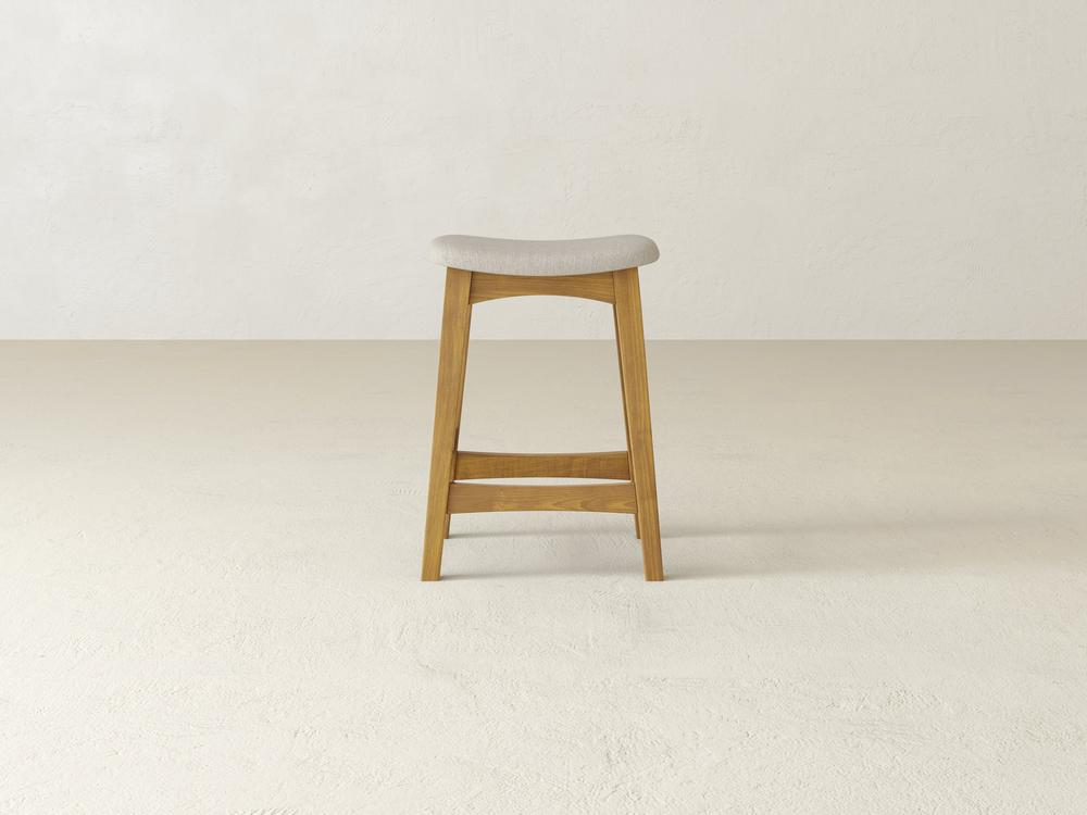 The Weston Upholstered Stool by James & James, crafted from durable hardwood, features a light gray cushioned seat, four sturdy legs, and a footrest. It stands against a matching light gray wall and floor, effortlessly enhancing the minimalistic and bright setting.