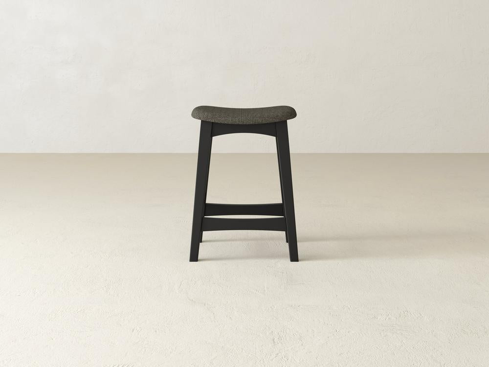 The James & James Weston Upholstered Stool, with a tall, minimalist design featuring a cushioned contoured seat, four legs, and a footrest, stands on a light textured floor against an off-white wall. Its dark gray color enhances its sleek appearance.