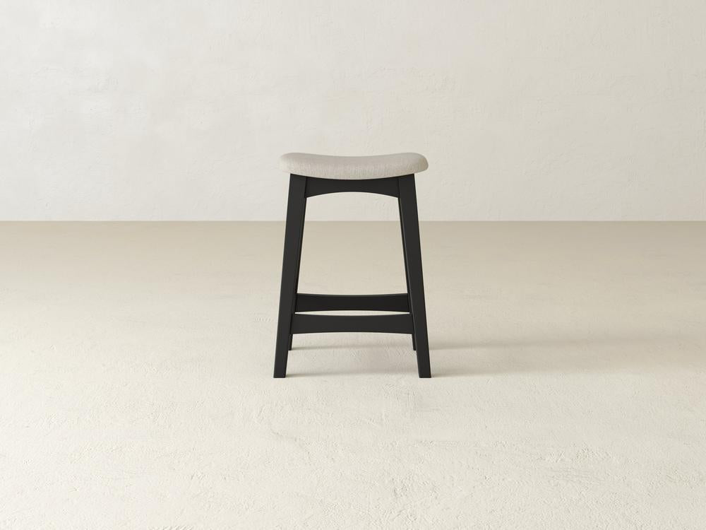 The Weston Upholstered Stool by James & James, featuring a minimalist design with a light gray cushioned seat and black maple hardwood frame, stands on a beige floor against a plain white wall.