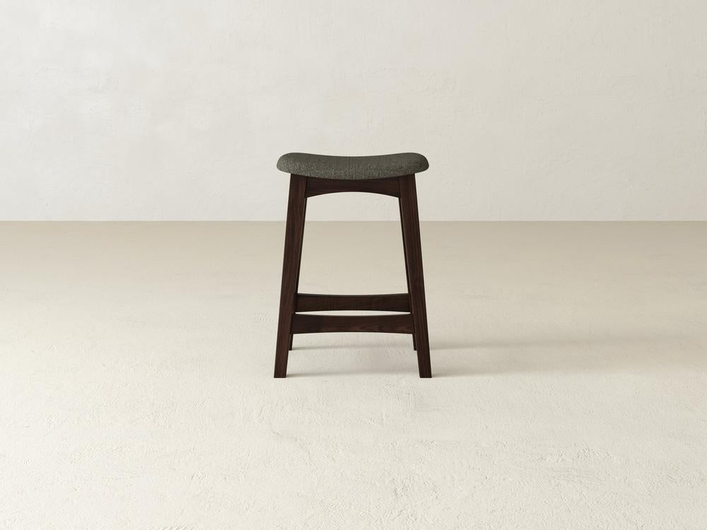 The James & James Weston Upholstered Stool, with a contoured gray fabric seat and tall dark maple hardwood frame, stands elegantly on a light beige floor against a white wall. Its minimalist design features tapered legs and a footrest, combining elegance and comfort.