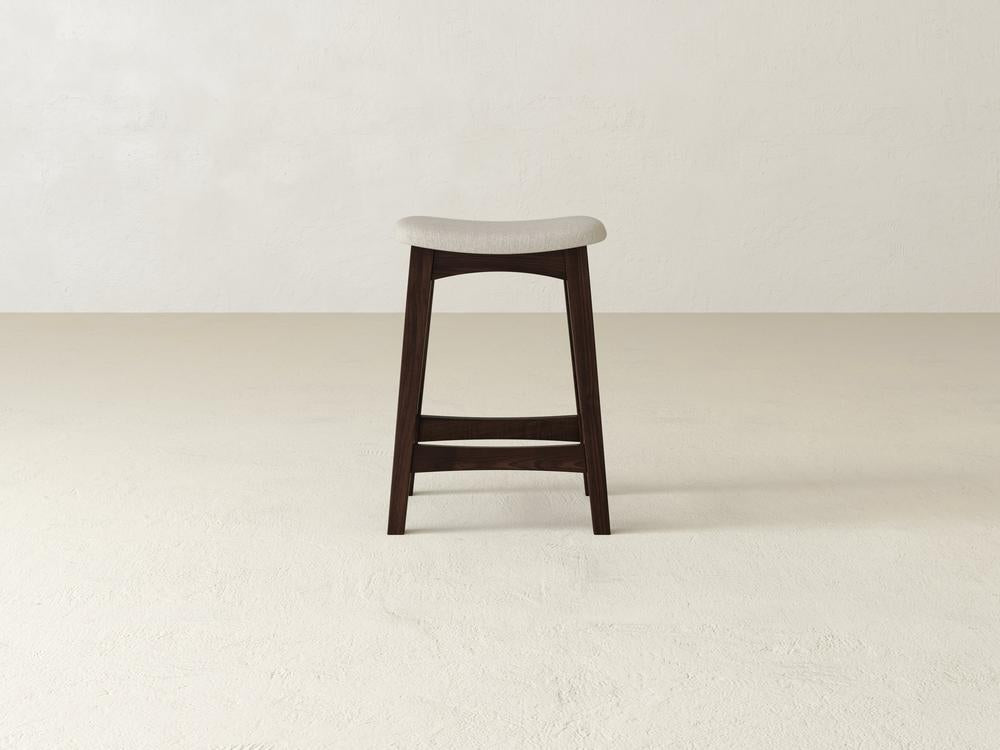 The James & James Weston Upholstered Stool showcases a minimalist design with a contoured light gray cushioned seat, durable maple hardwood dark legs, and an angular footrest, creating a sleek, modern look.
