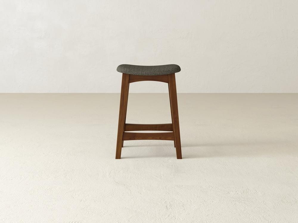 The Weston Upholstered Stool by James & James features a minimalist design with simple lines, a contoured gray cushioned seat, and a natural maple hardwood finish. It stands on a light beige floor against an off-white wall.