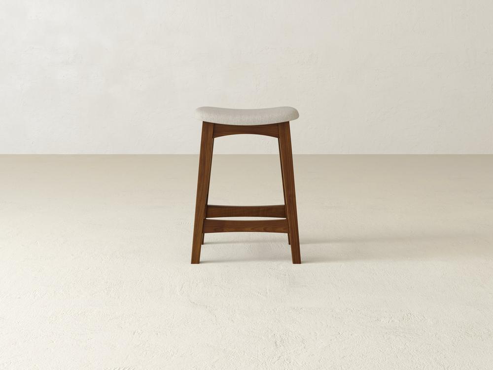 The Weston Upholstered Stool by James & James, featuring a curved beige cushion and crafted from durable maple hardwood, is centered in a minimalist room with light-colored walls and flooring.