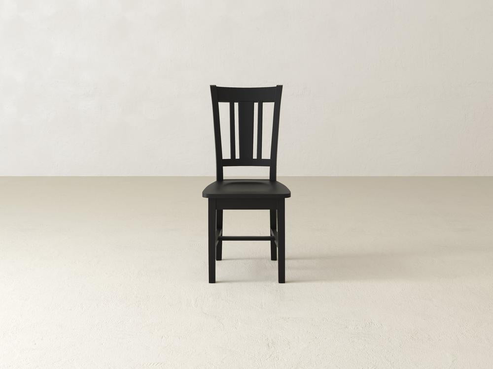 A wooden chair with a slatted backrest stands on a light-colored floor against a plain, off-white background.