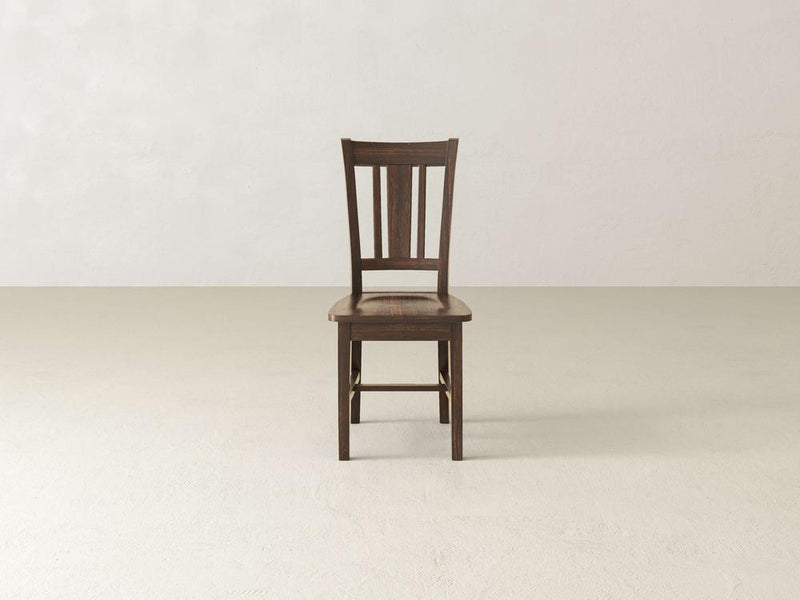 The William Dining Chair - Tobacco by James+James is centered in a minimalist room with light-colored walls and flooring. This dark wooden chair features a slatted backrest, four straight legs, and a simple yet classic design.