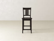 The William Stool by James & James, a handcrafted dark wooden seat, stands alone on a light floor against an off-white wall—ideal for minimalist home decor.