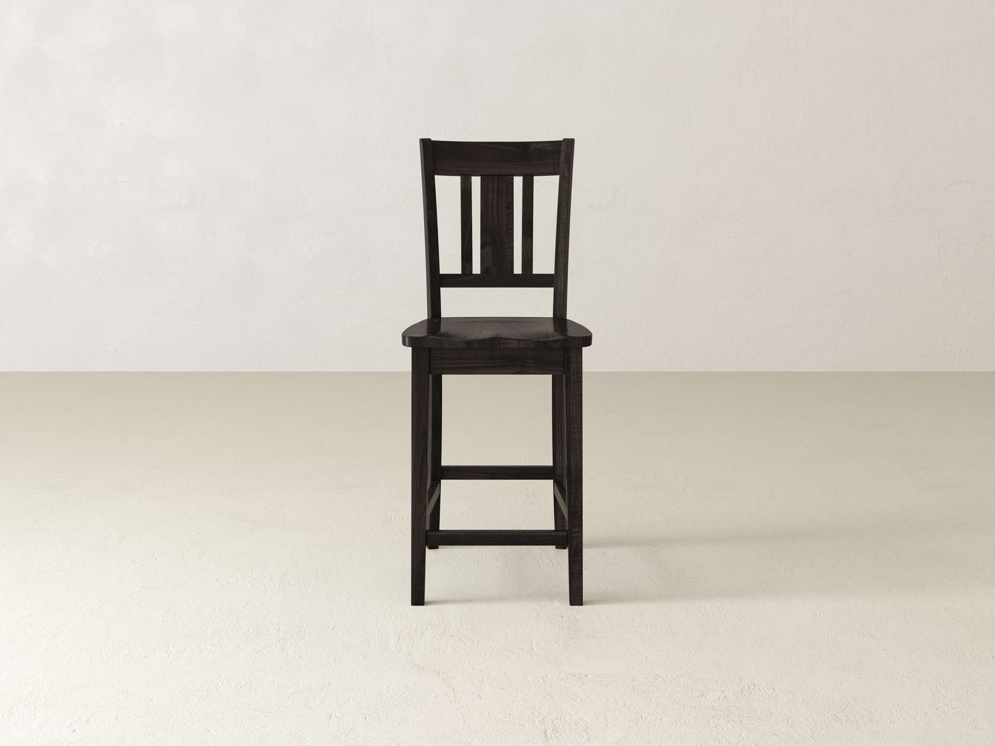 The William Stool by James & James, a handcrafted dark wooden seat, stands alone on a light floor against an off-white wall—ideal for minimalist home decor.