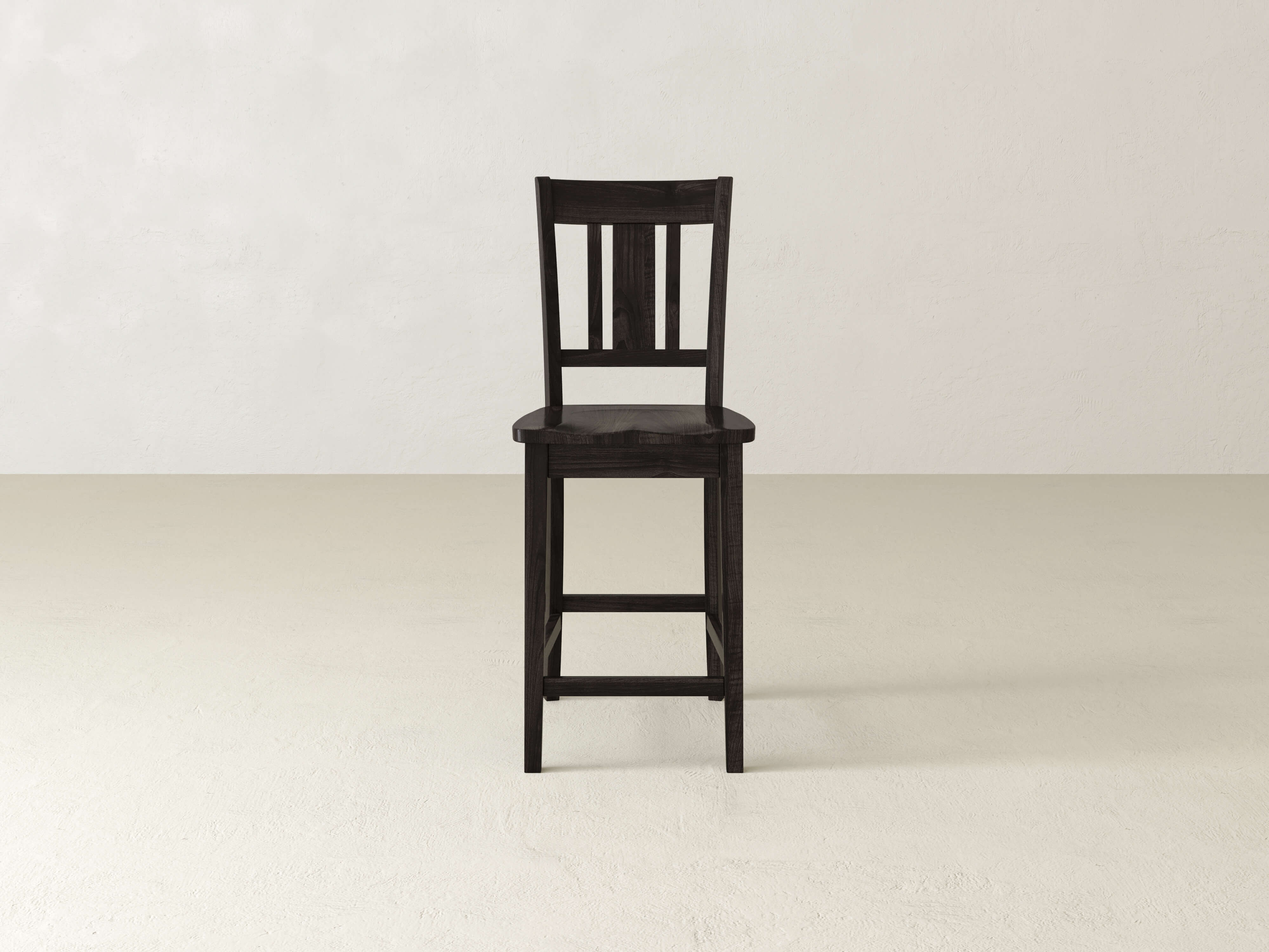 The William Stool by James & James, a handcrafted dark wooden seat, stands alone on a light floor against an off-white wall—ideal for minimalist home decor.