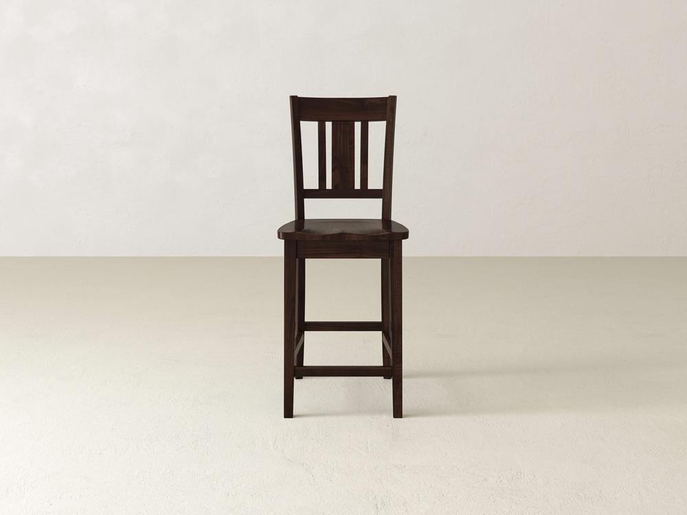 The William Stool by James & James exudes timeless sophistication on a plain light floor against an off-white wall. This elegant dark wooden piece features a slatted backrest and footrest, blending seamlessly into any home decor with its simple yet refined design.