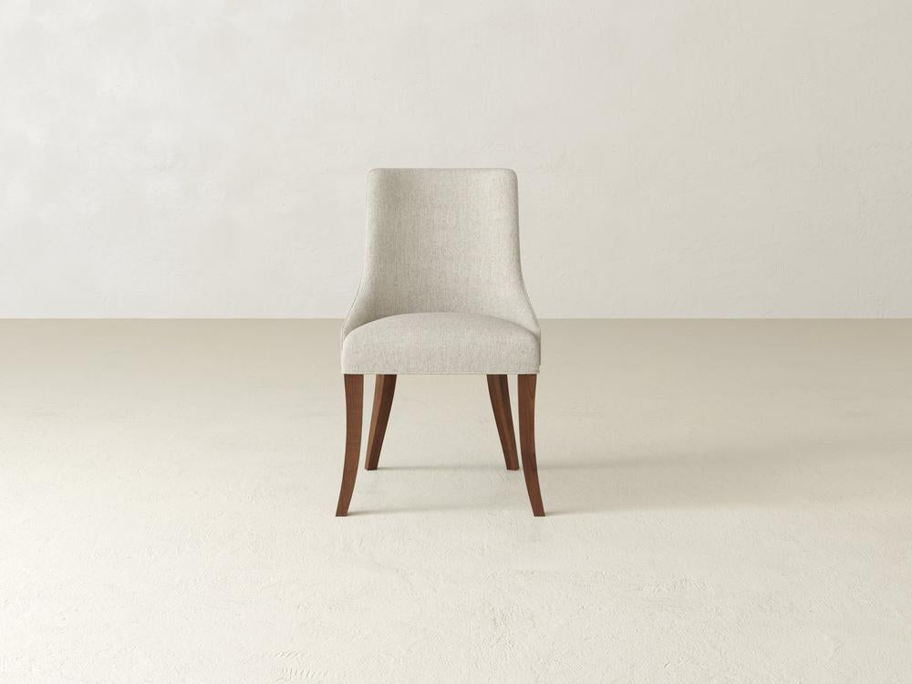 A minimalist, beige upholstered chair with wooden legs is centered in a plain, light-colored room. The design is simple and modern, with a slightly curved backrest and flared legs.
