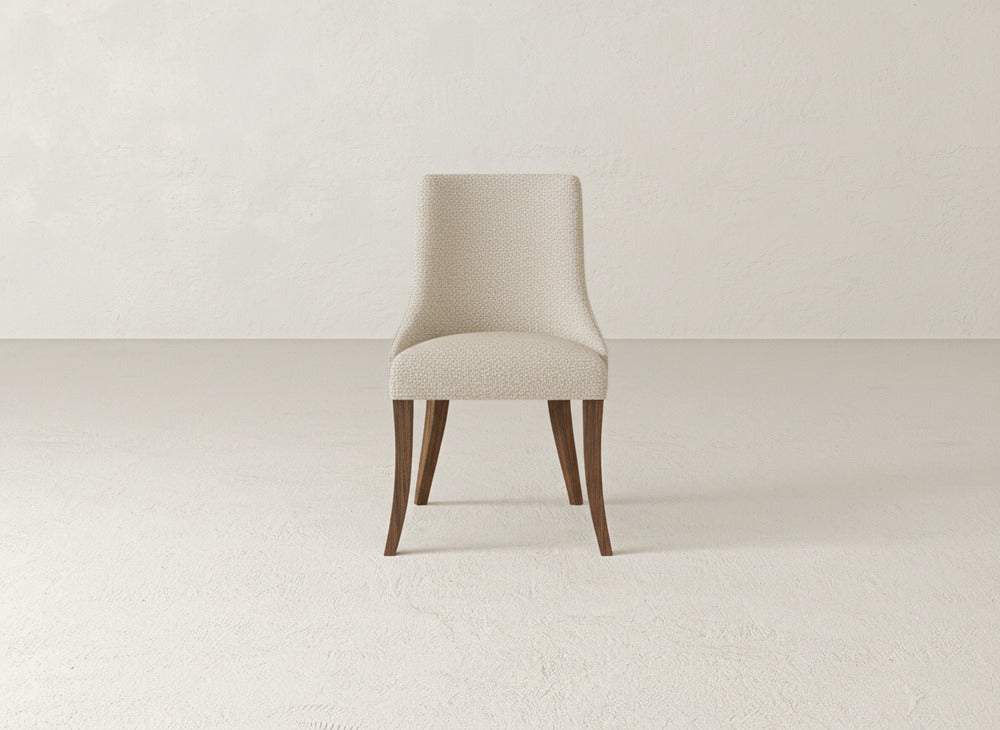 A modern James+James Willow Dining Chair, featuring a light beige upholstered seat and backrest with dark wooden legs, is centered in an empty minimalist room with off-white walls and flooring.