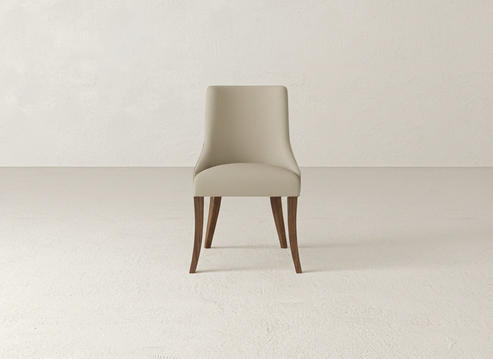The James+James Willow Dining Chair, with a light beige upholstered seat and backrest and four wooden legs, sits in a minimalist room with textured, light-colored walls and floor.