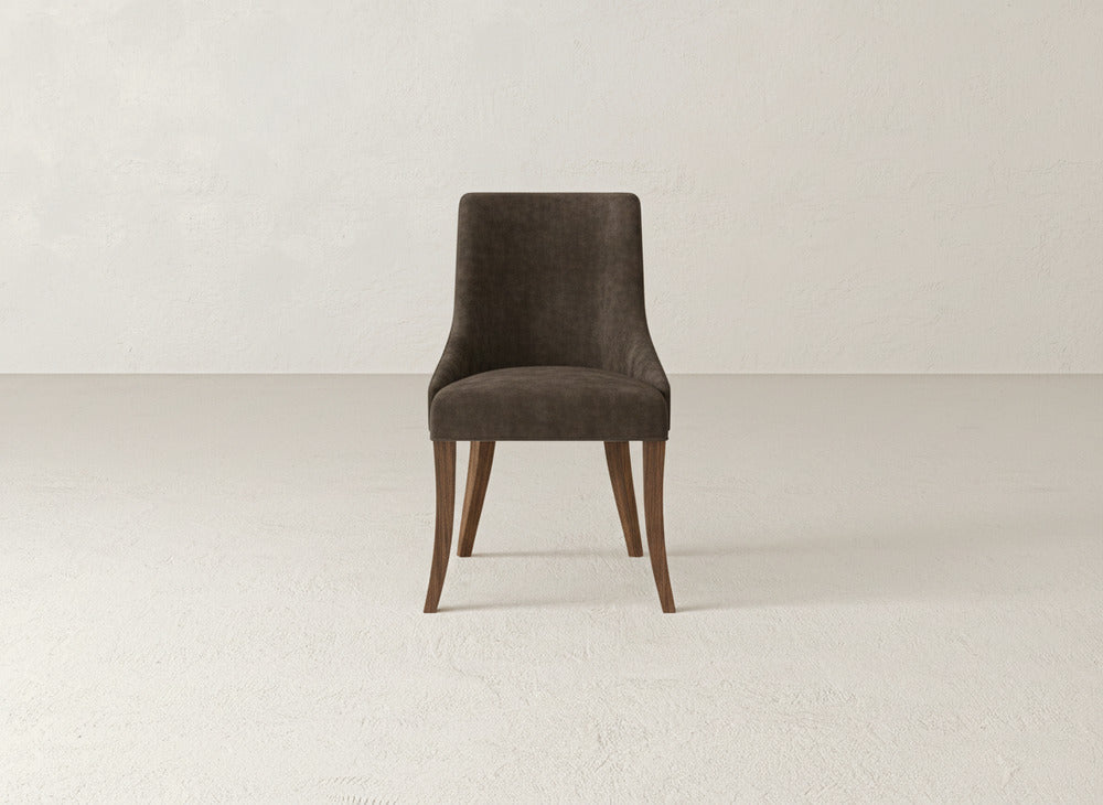 The Willow Dining Chair by James+James, with a dark upholstered seat and backrest and curved wooden legs, is centered in an empty, light-colored room featuring textured walls and floors.