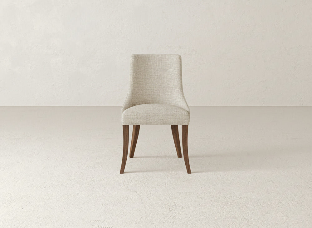 A James+James Willow Dining Chair with light beige upholstery and dark wooden legs is centered in a minimalist, neutral-toned room with a textured floor and plain wall.