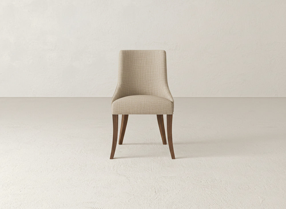 A beige James+James Willow Dining Chair with a curved backrest and wooden legs stands on a light-colored, textured floor against an off-white wall.