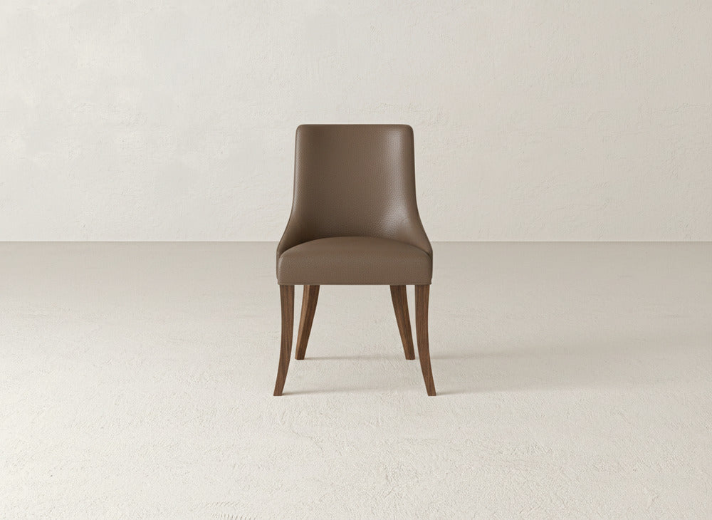 The Willow Dining Chair by James+James, featuring brown upholstery, a curved backrest, and wooden legs, is placed on a light floor against a textured white wall.