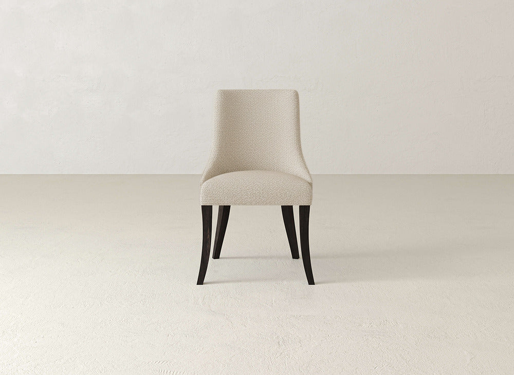 A single James+James Willow Dining Chair, featuring light beige upholstery and dark wood legs, sits centered in a minimalist room with light walls and floor.