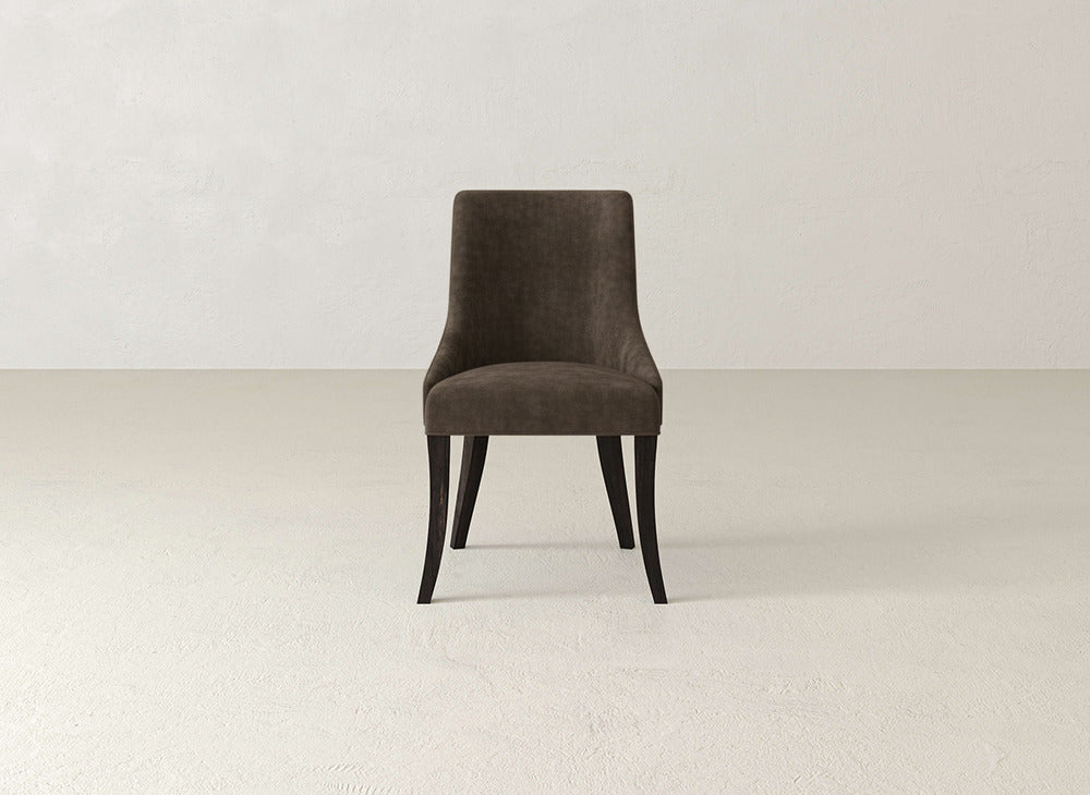 A James+James Willow Dining Chair with brown upholstery and dark wooden legs stands centered on a light textured floor, facing forward against a plain off-white wall in a minimalist setting.
