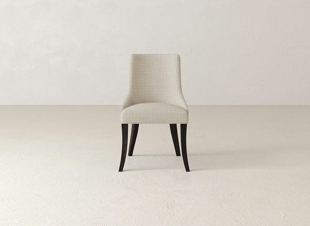 A James+James Willow Dining Chair with light beige fabric and dark wooden legs sits at the center of an empty, light-toned room with textured walls and floor.
