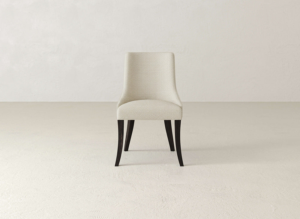 A beige James+James Willow Dining Chair with dark wood legs is centered in a minimalist room featuring light walls and flooring.