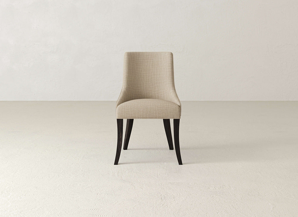 A beige James+James Willow Dining Chair with dark wood legs is centered in an empty, light room with plain walls and a smooth floor.