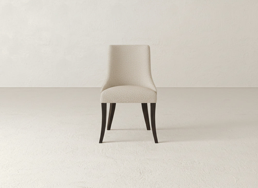 The Willow Dining Chair by James+James, featuring modern beige upholstery and dark wooden legs, is placed at the center of an empty, light-toned room with textured walls and flooring.