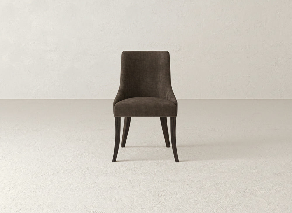 The Willow Dining Chair by James+James, upholstered in dark brown with curved wooden legs, rests on a light textured floor against a plain off-white wall, enhancing the minimalistic and uncluttered setting.