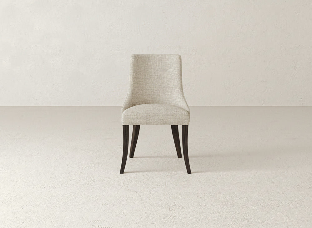 The James+James Willow Dining Chair, upholstered in light beige with dark wooden legs, is centrally placed against a textured off-white wall and floor background.