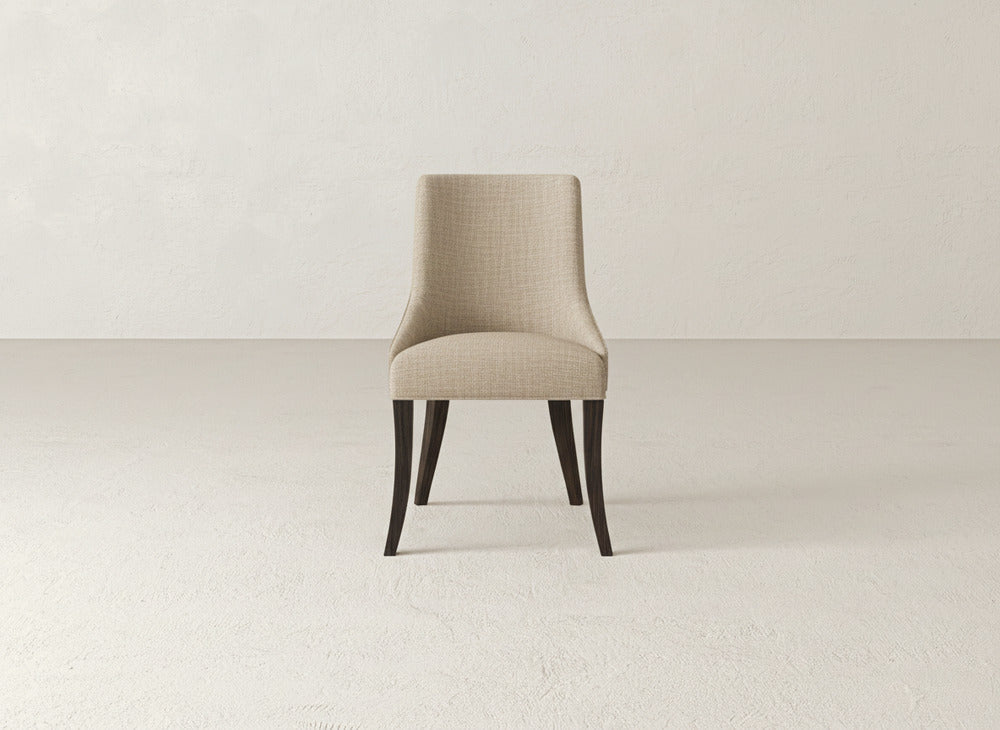 A beige James+James Willow Dining Chair with dark wooden legs sits at the center of a minimalist room featuring a light, textured wall and floor.