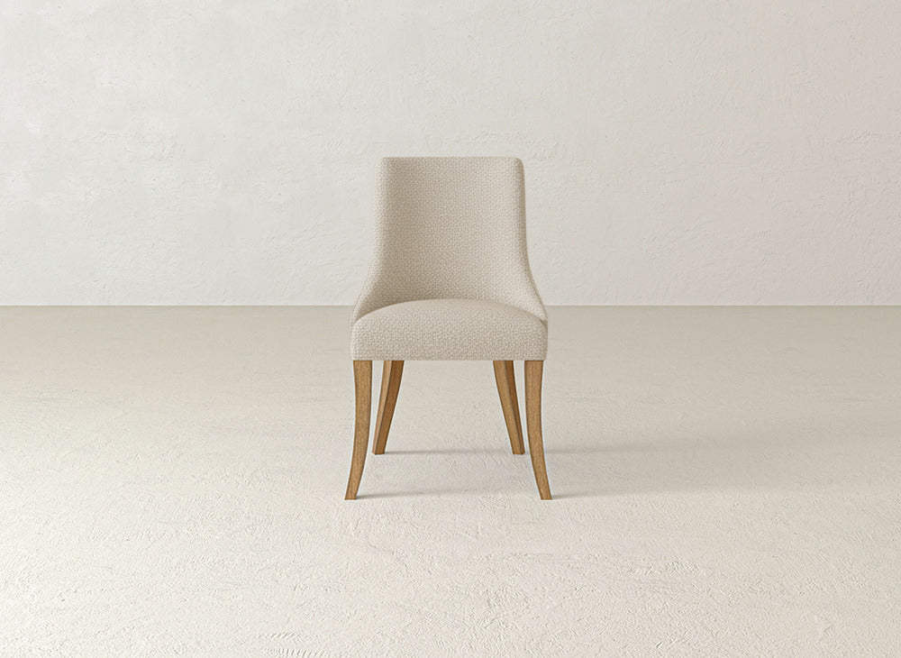 The Willow Dining Chair by James+James, featuring beige upholstery and wooden legs, stands in the center of a light-colored room with plain walls and a textured floor.
