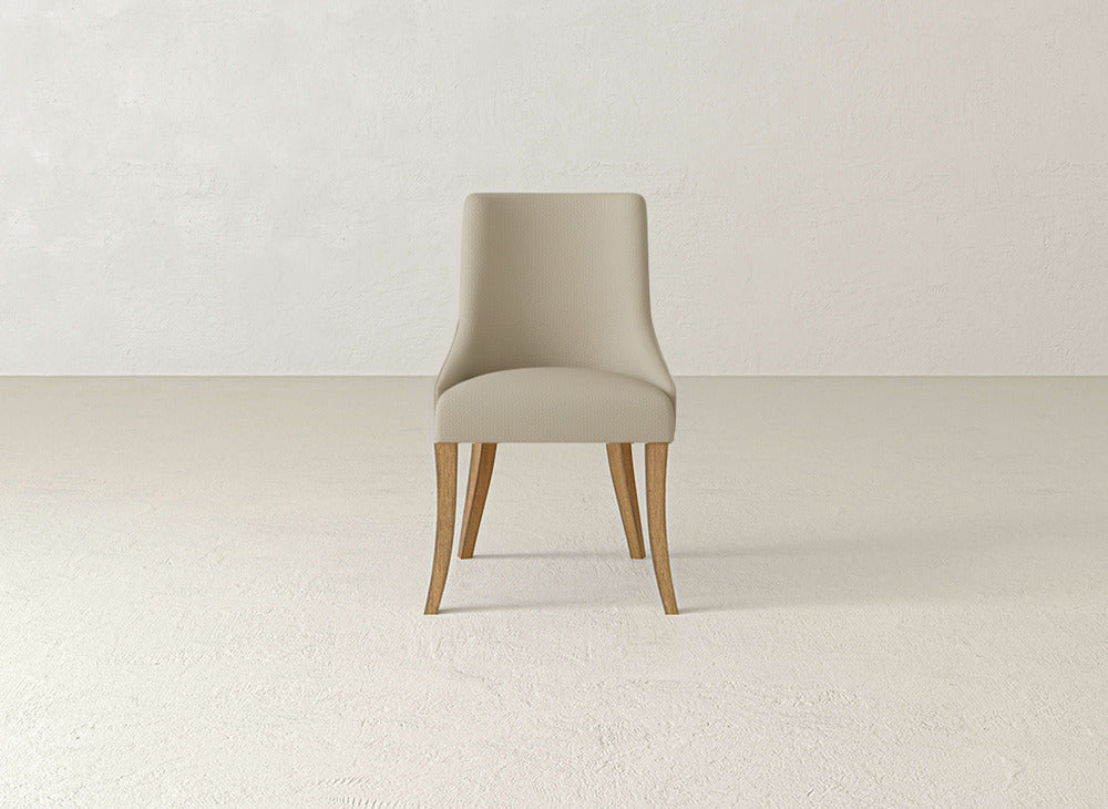 The Willow Dining Chair by James+James, featuring light beige upholstery and wooden legs, is centered in a minimal, light-toned room with a textured floor and plain wall.