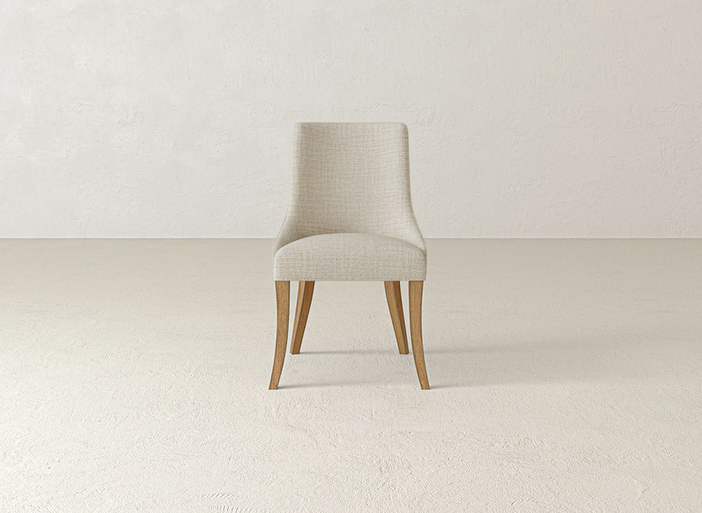 The James+James Willow Dining Chair features light beige upholstery and four wooden legs, adding modern elegance to a minimalist room with a light textured floor and wall.
