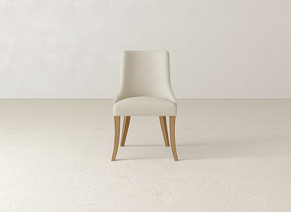 A James+James Willow Dining Chair featuring beige upholstery and wooden legs is centered in a minimal, empty room with off-white textured walls and flooring.
