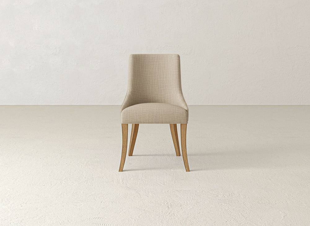 A beige James+James Willow Dining Chair with wooden legs sits centered in an empty, light-toned room with textured walls and floors.
