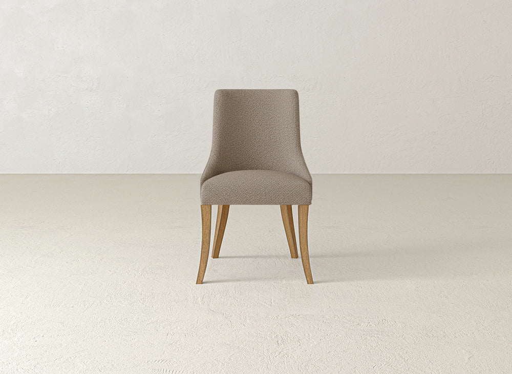A James+James Willow Dining Chair with light brown fabric and wooden legs is placed on a plain, light floor against a textured off-white wall.