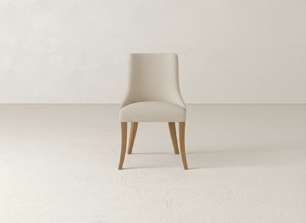 A James+James Willow Dining Chair with beige upholstery and light wooden legs is centered in a neutral-toned room featuring a textured wall and floor.