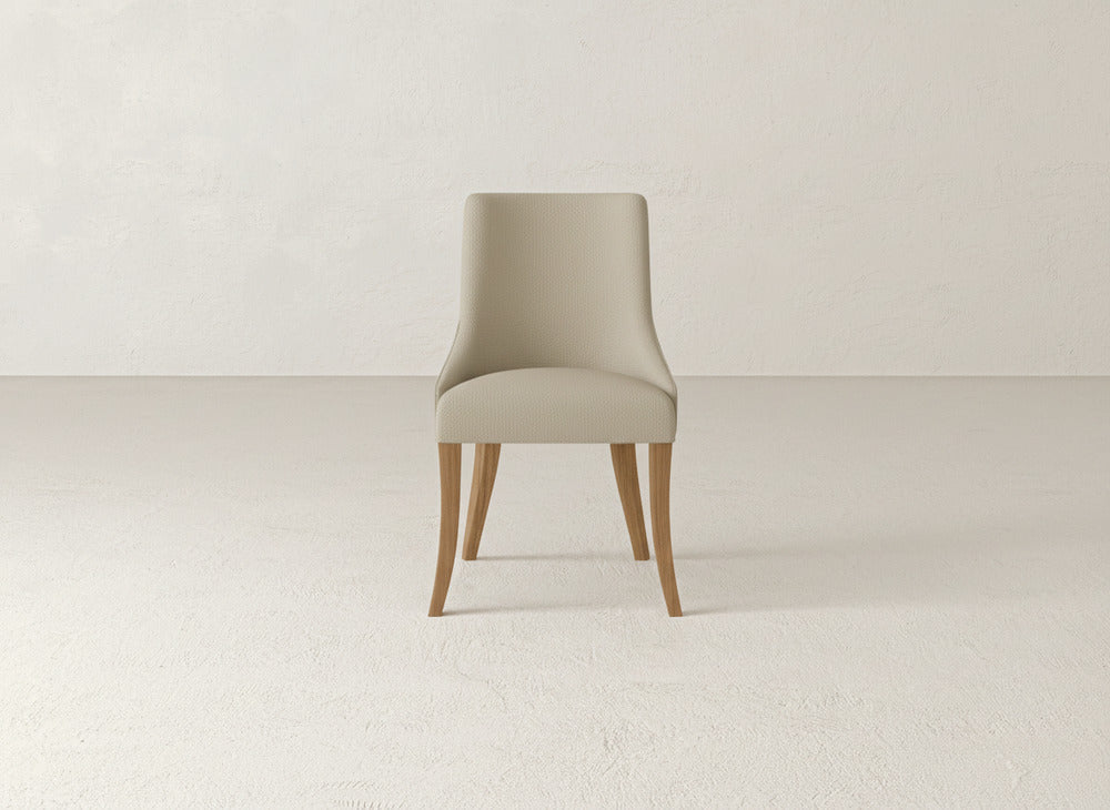 A James+James Willow Dining Chair with light beige upholstery and wooden legs sits centered in an empty, light-toned room featuring textured walls and flooring.