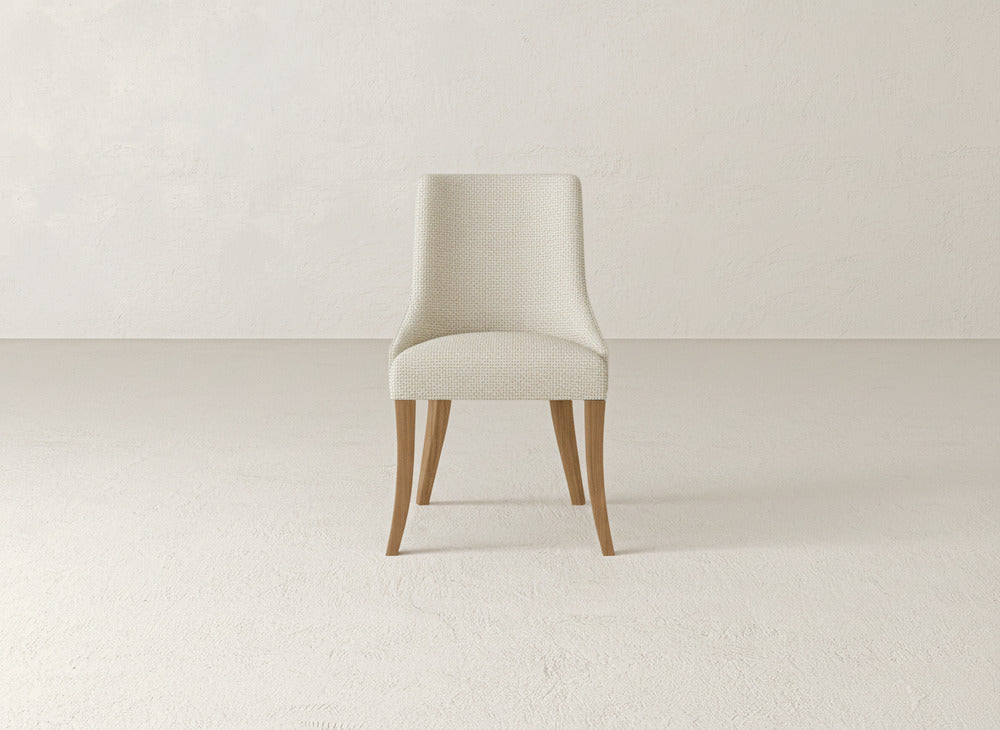 The James+James Willow Dining Chair, featuring light beige upholstery and wooden legs, is centrally placed against a plain, light-colored wall and floor.