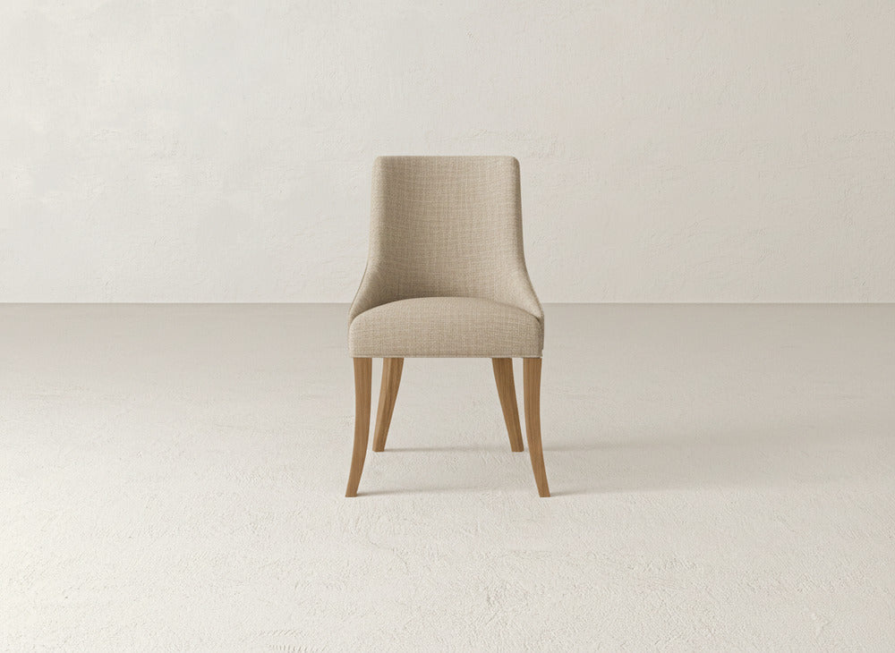 A beige James+James Willow Dining Chair with light wooden legs sits centered in a minimal, empty room featuring a light-textured floor and plain walls.