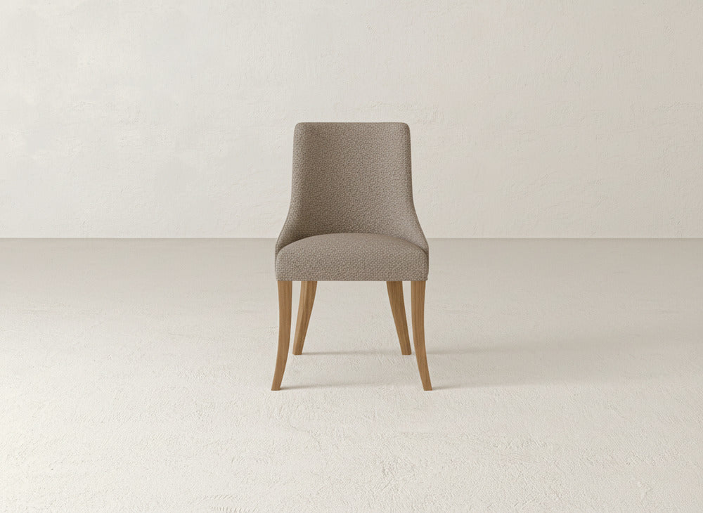 The Willow Dining Chair by James+James features a modern design with a curved back and seat in light beige upholstery, supported by four angled wooden legs, shown against a simple, light background.