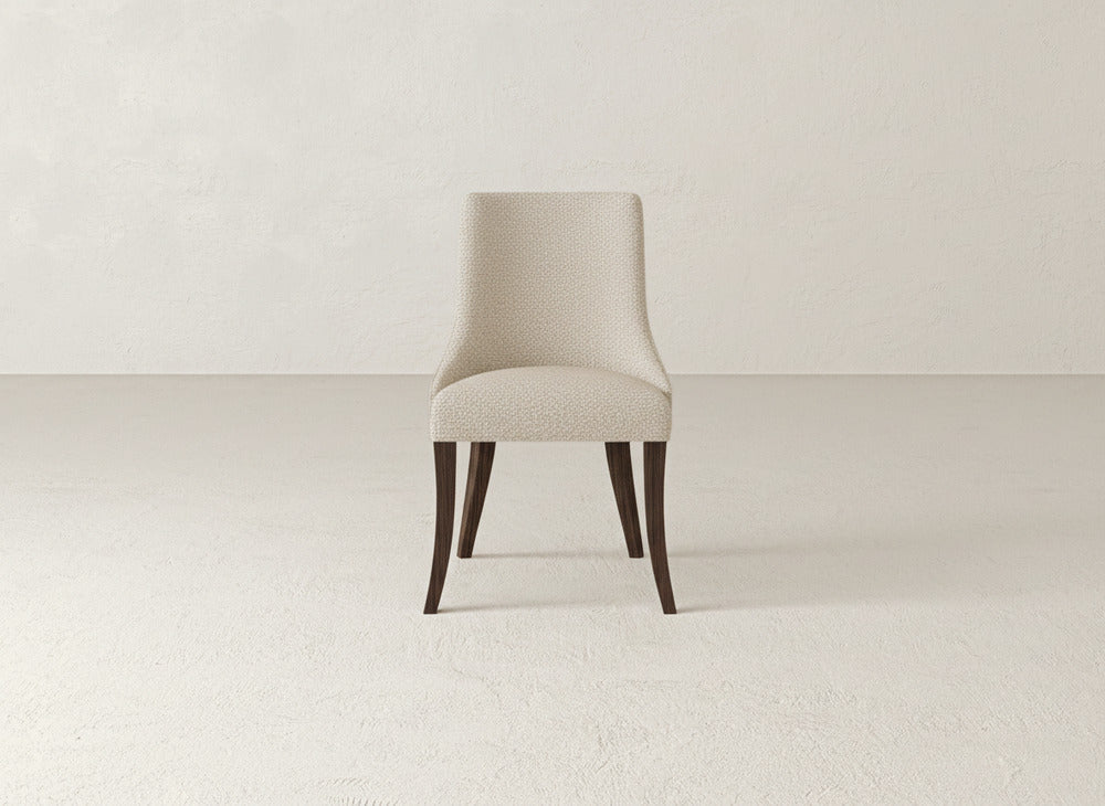A James+James Willow Dining Chair with light beige upholstery and dark wooden legs is centered in an empty, neutral-toned room with textured walls and floors.