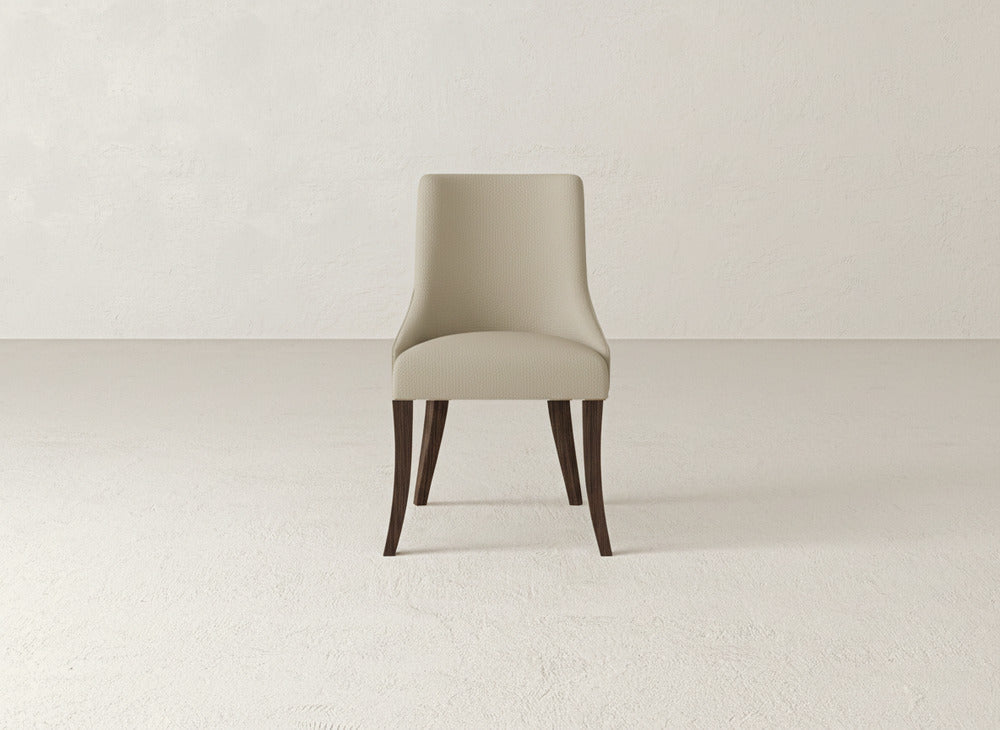 The Willow Dining Chair by James+James features modern beige upholstery and dark wooden legs, offering understated elegance to any space when placed in a room with light-colored textured walls and floors.