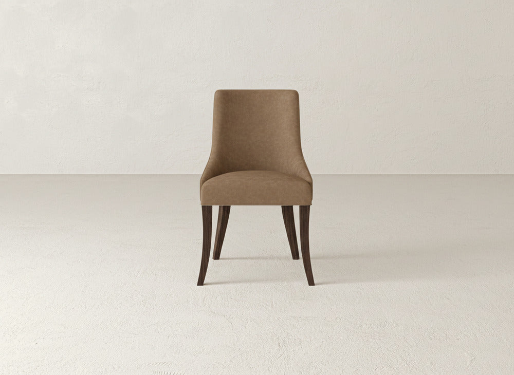 The Willow Dining Chair by James+James, featuring brown upholstery and dark wooden legs, is centered in a minimalist, light-toned room with a simple textured wall and floor.