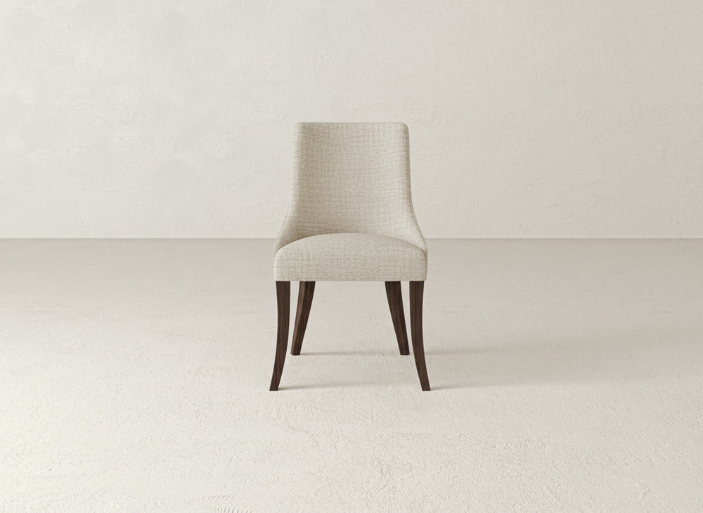 The James+James Willow Dining Chair features modern beige upholstery, a curved backrest, and dark wooden legs, showcased in the center of an empty, light-colored room with a textured wall and floor.