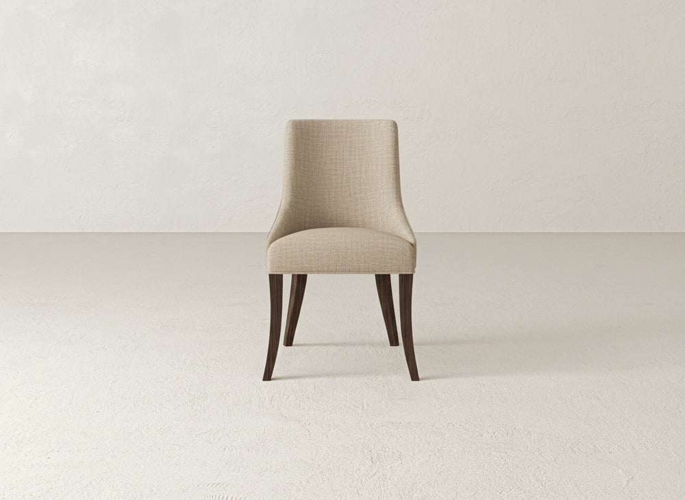 The Willow Dining Chair by James+James, upholstered in beige with dark wooden legs, is placed at the center of a minimalist, light-toned room with a plain textured wall and floor.
